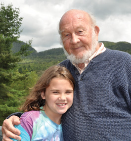 Man with a beard poses with a little girl, with mountains, trees and clouds in the background